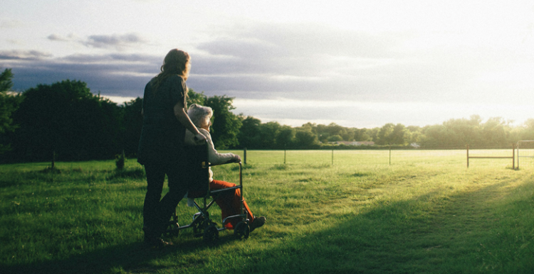 Photo of a middle aged woman pushing an older woman in a wheelchair across a field. It looks like early morning light in the sky, the sun is low on the horizon