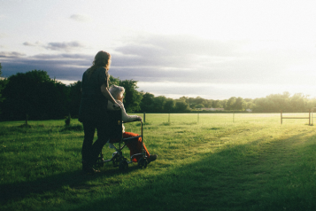 Photo of a middle aged woman pushing an older woman in a wheelchair across a field. It looks like early morning light in the sky, the sun is low on the horizon