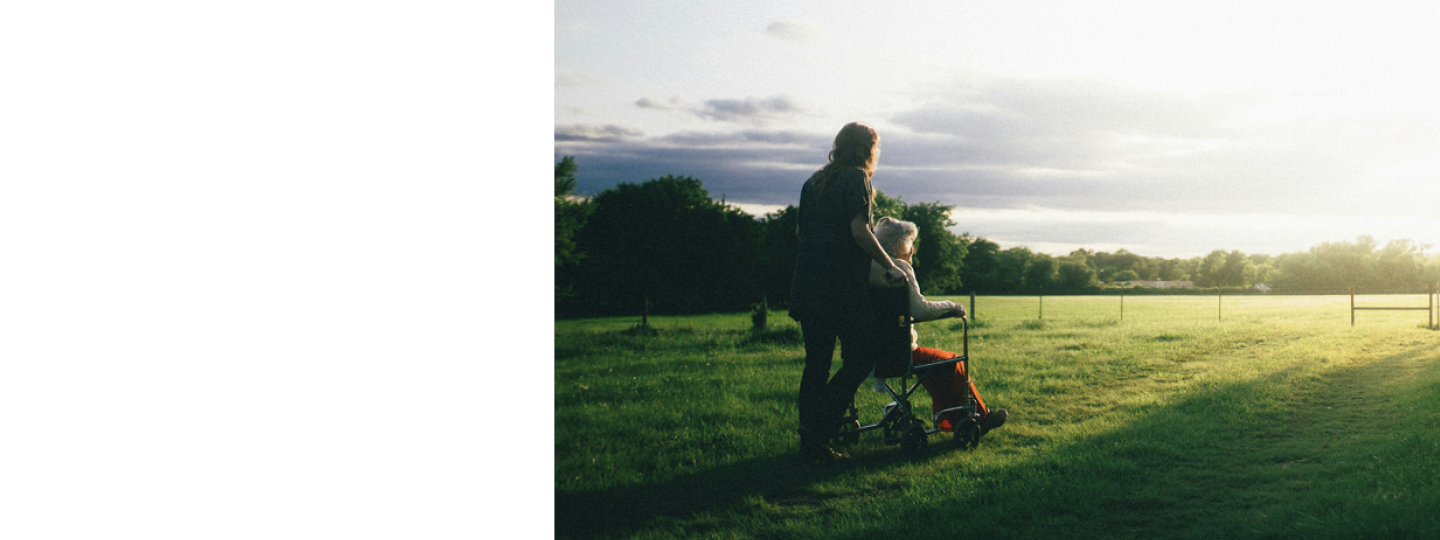 Photo of a middle aged woman pushing an older woman in a wheelchair across a field. It looks like early morning light in the sky, the sun is low on the horizon