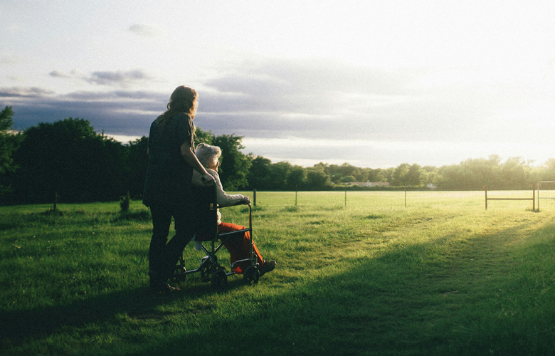 Photo of a middle aged woman pushing an older woman in a wheelchair across a field. It looks like early morning light in the sky, the sun is low on the horizon